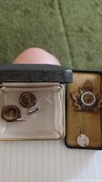 Photo of two cufflinks with painted portraits in a vintage box on left, WWI battalion pin and medallion in vintage case on right