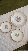 Three vintage plates on a white quilted surface, the largest has a pink rose with beige gold border, two smaller with floral designs and gold rims.