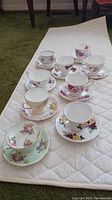 Nine vintage English teacups and saucers arranged on a white quilted mat. Various floral patterns and color schemes visible.