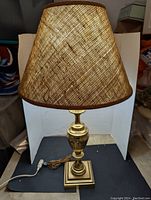 Front view of brass table lamp with shade on, showing woven fabric shade and polished brass base with ribbed urn design.