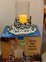 Full view of Christmas village candle holder on original box, showing snowy village base with candle inside glass hurricane