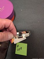 Hand holding a silver-tone bracelet with visible engraved text and a Kate Spade tag next to a purple and orange Kate Spade branded box.