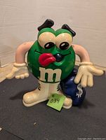 Front view of green M&M character piggy bank showing facial expression, body with white 'm', white boots and gloves, and money bag in the left hand.