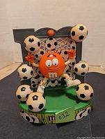 Front view of the orange M&M character soccer themed dispenser with soccer balls around and green base.