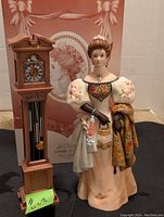 Front view of porcelain Mrs. Albee figurine standing next to grandfather clock, with original box in background