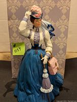 Front view of the porcelain figurine showing a Victorian woman in detailed clothing holding a purse and adjusting her feathered hat.