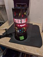 Front photo of cylindrical LED bar light sign showing 'THE BAR is open' text illuminated with multi-colored light patterns on a black base with cord.