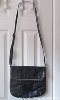 Black faux leather crossbody bag hanging on a white door, showing the front and strap.