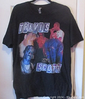 Front view of black Travis Scott T-shirt on hanger showing graphic design with text and images of Travis Scott.