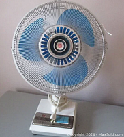 Front right side view of vintage oscillating table fan showing control panel, blue blades, and white base with plug wrapped around stand.