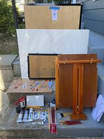 Photo of wooden easel, two wooden watercolor paper stretchers, two large watercolor papers, a palette board with paint splotches, assorted paint tubes, brushes, and pencils arranged on steps outdoors.