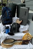 Complete camping gear set arranged outdoors on a white cloth including sleeping bags, backpack, sleeping mat, water container, fireplace toasters, tennis rackets, and a baseball glove.