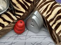Close-up of Kong red rubber dog toy and Modus anti-bark device with silver triangle shape, placed on grey and zebra-patterned fabric.
