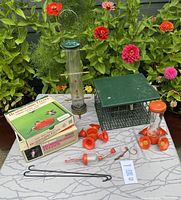Photo showing all three feeders with box for one hummingbird feeder and extra parts laid on table with flowers in background.