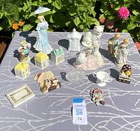 Photo of assortment of vintage china and porcelain figurines displayed outdoors on a patterned gray cloth, showing various figures including women, children, cats, birds, houses, and decorative items.