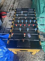 Foosball table shown at an angle, displaying playing surface with red and blue men on rods and black handles visible.