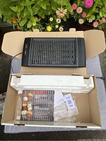 Top down view of the black electric indoor grill in open box showing the grill plate and packaging contents like manual and papers.