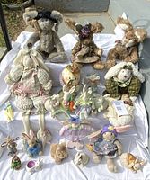 Photo showing collection of bunny plush toys and figurines arranged on white cloth outdoors. Includes three large plush bunnies, one musical bunny with basket and purple hat, several smaller bunny figurines and dolls in pastel colors