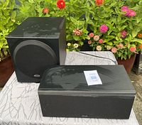 Polk Audio subwoofer and center speaker displayed on a cloth-covered table with garden background.