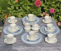 Photo of tea set with cups on saucers, dinner and side plates, cream and sugar set arranged on tablecloth in garden setting.
