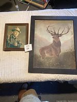 Two framed art pieces on a bed, one portrait photo of a man and one elk artwork