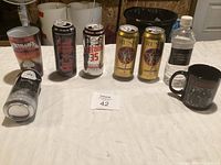 Metallica tumbler, AC/DC tumbler, four beer cans including two Rush brand and one Rye Lager can, a black mug, and a water bottle on a table.