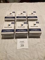 6 boxes of Pencil Buddies triangle shaped golf pencils, stacked on a white surface with a lot tag number 39 visible.