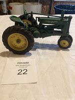 Side view of green John Deere Model G metal tractor showing large yellow rear wheel and smaller front wheel, steering wheel, and exhaust pipe.