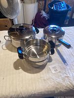 Overview of all three parts of the set placed on a surface including large pot with lid, smaller pot with lid and long handle, and steamer insert.