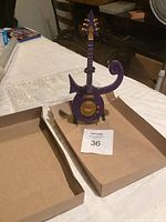 Miniature purple electric guitar on matching purple stand, displayed on white surface with open box