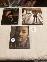 Three musician books laid out on a bed: Leonard Cohen Everybody Knows, On Tour by Dave Marsh, and Bruce Springsteen Songs.
