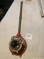 Full view of the pipe threader showing the tool with a long handle and red base, placed on a white surface along with a paper label showing number 70.