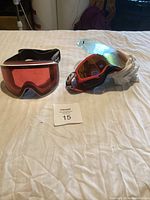 Two pairs of ski and snowboarding goggles on a white surface with a lot number card; one pair has a red lens with a black and white frame, the other with a red lens and white strap.