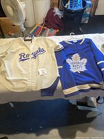 Photo showing white Royals baseball jersey with blue script logo and blue Toronto Maple Leafs hockey jersey with white maple leaf logo displayed sideways on a table.