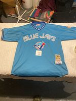 Full view of light blue Toronto Blue Jays jersey with large logo and 'Blue Jays' text on chest laid flat on white surface.