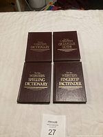 Four New Webster's books laid out on a white cloth surface, showing covers labeled Dictionary, Grammar Guide, Spelling Dictionary, and Fingertip Factfinder.