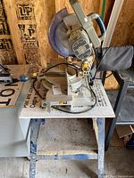 Front angle photo of the 10-inch compound mitre saw on metal framed table stand, showing saw body, handle, blade guard, and dust bag.