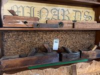 Photo of a wooden shelf holding two antique wood spirit levels on the top shelf and four antique wooden hand planers on the lower shelf, showing general view of all tools.