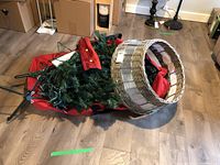 Christmas tree components including the tree, metal stand, red canvas carrying bag, and wicker base cover laid on floor to show all included items.