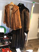 Both fur coats hanging side by side on hangers showing full length and relationship.