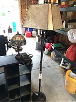 Photo showing both lamps: bronze floor lamp with tri-light parchment shade and ornate stained glass table lamp on a black shelf in a garage setting.