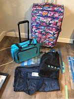Five bags displayed on floor: large rolling suitcase with colorful print and handle, small turquoise Air Canada rolling bag, black Smart Set bag, Impala garment bag, dark blue carry bag