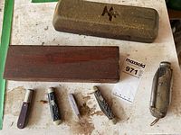 Lot showing four pocket knives in front of two sharpening stones, one with MF mark and wooden box; the other sharpening stone lying closed in wooden box