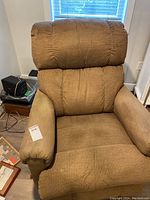 Front view of the light brown chenille rocker recliner showing padded arms and cushioned back with visible wear on the armrest.