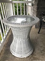 Side view of round white wicker table on porch floor with glass inset top and foliage visible behind railing.