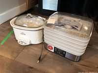 White Rival Crock-Pot slow cooker with glass lid and Hamilton Beach food dehydrator with clear plastic trays and stainless steel base.