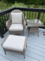 Full set of an outdoor patio chair with arms and cushions, matching ottoman with cushion, and square side table with glass top on a wooden deck.