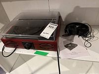 Left angled view showing Pyle turntable with clear dust cover, wood finish, front speakers and volume knob, black power cord visible. Sony headphones and user manual are to right of turntable.