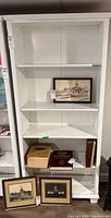 Front view of white wooden bookcase showing five shelves with some books and framed pictures placed on it for staging.