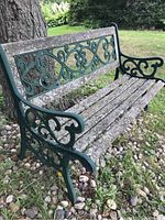 Front left angled view of green painted wrought iron garden bench showing ornate scrollwork and weathered wooden slats with signs of wood rot.